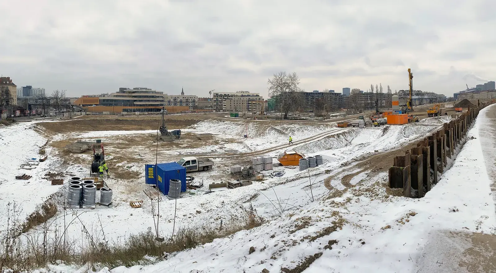 Weite Baugrube im Winter, am Rand eine Spundwand, im Hintergrund Bohrgeräte und die Stadt